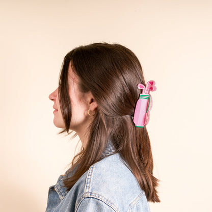 A woman with brown hair in profile wears a denim jacket and styles her hair with The Darling Effect’s Golf Clubs Shape Up Claw Clip—a pink, golf bag–shaped accessory—against a plain, light background.