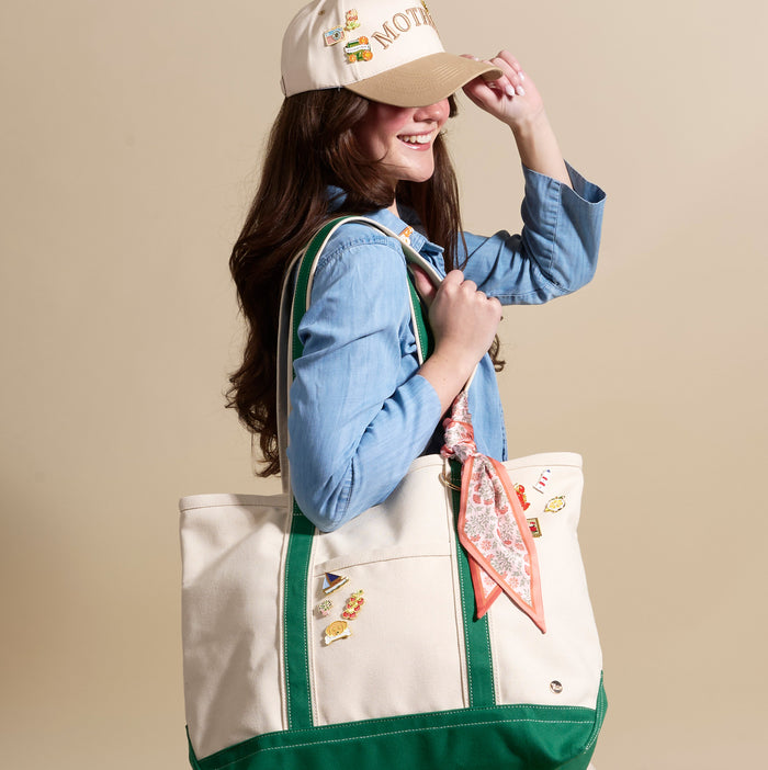 A smiling woman in a denim shirt and a Mother baseball cap holds a cream tote with green accents, decorated with The Darling Effect’s Golf Par-Tee The Pin Pack and a floral scarf.