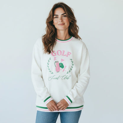 A woman with wavy brown hair wears The Darling Effect's Golf Social Club Oversized Crew, featuring green and pink accents, GOLF text above a golf graphic, and embroidered designs. She smiles slightly against a plain white background.