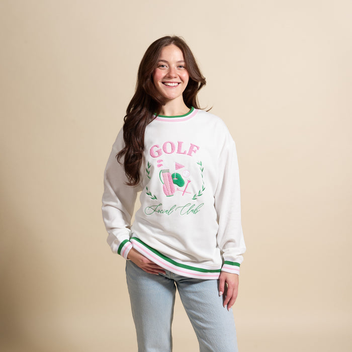 A young woman with long brown hair smiles against a beige background, wearing The Darling Effect’s Golf Social Club Oversized Crew—an oversized white sweatshirt with green and pink trim—and light blue jeans, one hand in her pocket.