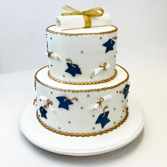 The Graduation Tiered Cake is iced in white buttercream and decorated with blue fondant grad caps and scrolls along the sides, plus a fondant scroll and gold ribbon on top.