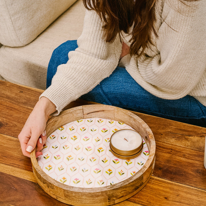 A person in a cream sweater and blue jeans sits on a wooden floor, holding the-darling-effect Handpicked For You Large Round Wood Enamel Tray with a floral pattern and a white candle on it.