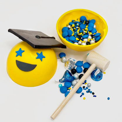 An opened up chocolate happy face grad showing the assorted candies inside with a mallet. The happy face is yellow and has blue stars for eyes, a smiling mouth, and a cap. A ballot is included to smash this piñata.