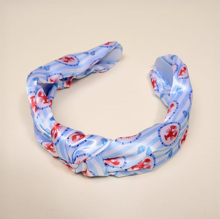 The Darling Effect's Harbor House Classic Twist Headband showcases a light blue fabric with red crab and blue shell patterns, accented by a stylish knot. Displayed on a beige background, this headband is a versatile accessory for any occasion.