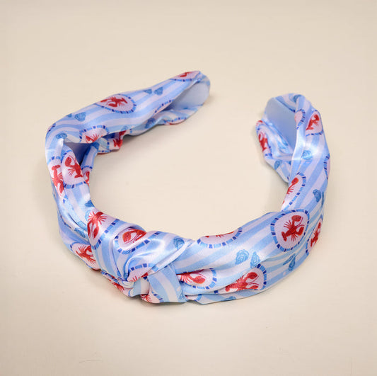 The Darling Effect's Harbor House Classic Twist Headband showcases a light blue fabric with red crab and blue shell patterns, accented by a stylish knot. Displayed on a beige background, this headband is a versatile accessory for any occasion.