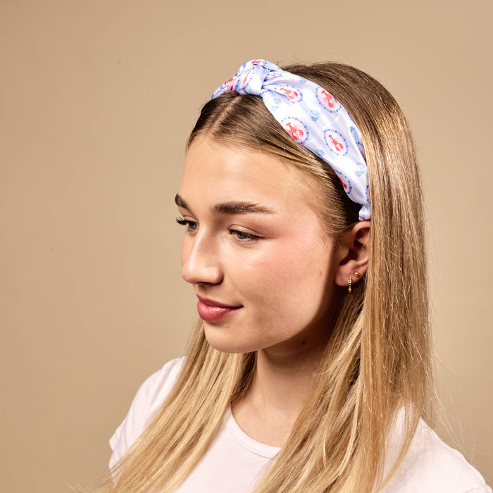 A young woman with straight blonde hair wears The Darling Effect's Harbor House Classic Twist Headband, featuring a blue and red circular pattern, paired with a white t-shirt against a beige background. She looks slightly downward and smiles softly.