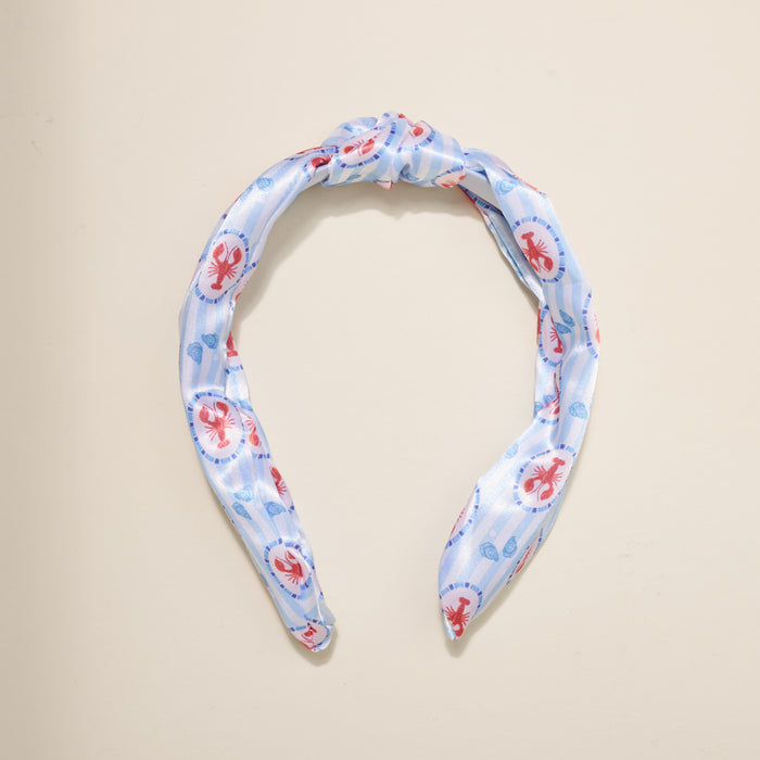 The Darling Effect’s Harbor House Classic Twist Headband features a light blue fabric with red lobsters and blue seashells, styled in an elegant top knot and shown on a beige background—a playful coastal accessory for any outfit.