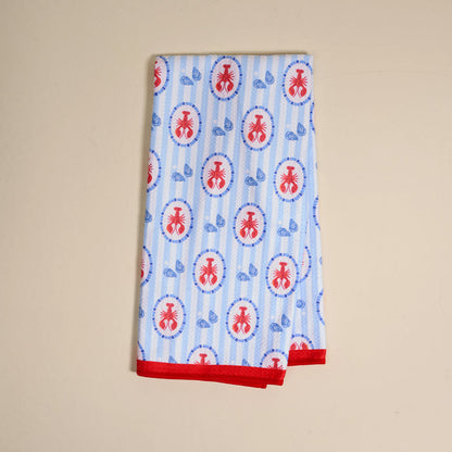 The Harbor House Waffle Tea Towel by The Darling Effect features a folded white waffle texture with blue stripes, red ant ovals, small blue leaves, and red-trimmed edges, shown on a beige surface.
