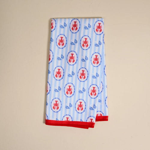 The Harbor House Waffle Tea Towel by The Darling Effect features a folded white waffle texture with blue stripes, red ant ovals, small blue leaves, and red-trimmed edges, shown on a beige surface.