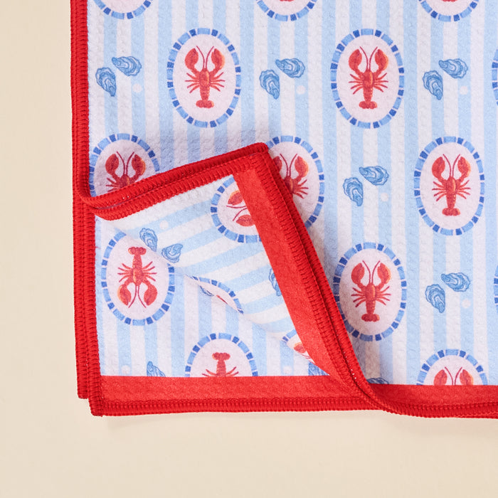 The Darling Effect Harbor House Waffle Tea Towel features a red border, blue stripes, and repeating red lobster and blue shell patterns. One corner is folded to display the underside of this absorbent kitchen towel.