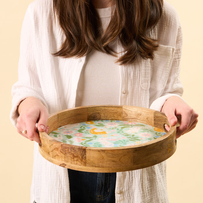 A woman in a white shirt and jeans holds the-darling-effect's Hoppy Days Large Round Wood Enamel Tray, which showcases a vibrant floral and animal design at its center, set against a light beige background.