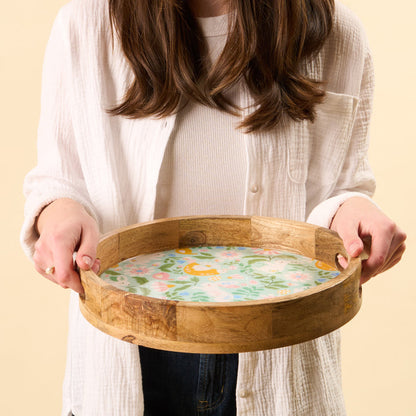 A woman in a white shirt and jeans holds the-darling-effect's Hoppy Days Large Round Wood Enamel Tray, which showcases a vibrant floral and animal design at its center, set against a light beige background.