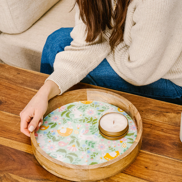 A person in a cream sweater and blue jeans sits at a wooden table, holding the-darling-effect's Hoppy Days Large Round Wood Enamel Tray with a colorful floral pattern and a stack of round coasters on it.
