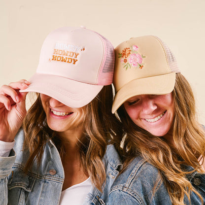 Two women in denim jackets and trucker hats from the-darling-effect smile together. One wears the pink "Howdy Howdy Howdy Trucker Hat"; the other sports a beige floral-embroidered hat. Both tilt their hats playfully.