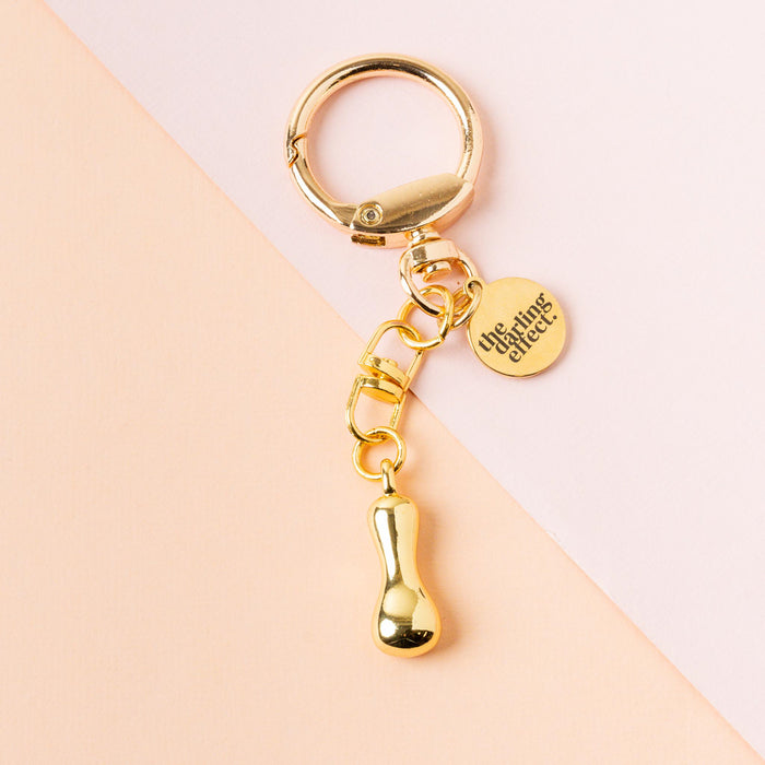 The I Bubble Letter Keychain Bag Charm from the-darling-effect features a gold keychain with a small round engraved tag and a shiny droplet charm, displayed on a soft pink and peach background.