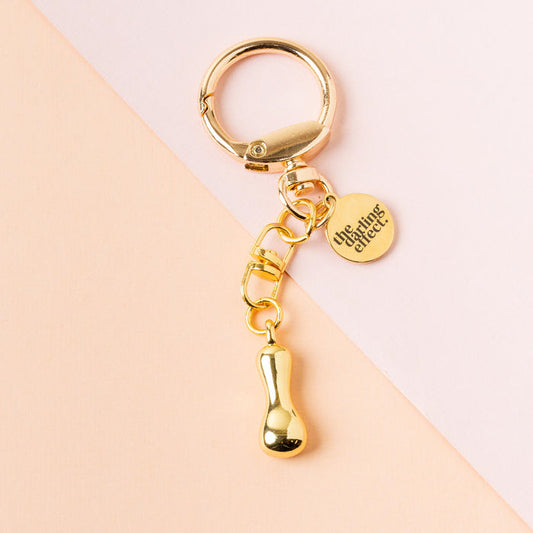 The I Bubble Letter Keychain Bag Charm from the-darling-effect features a gold keychain with a small round engraved tag and a shiny droplet charm, displayed on a soft pink and peach background.