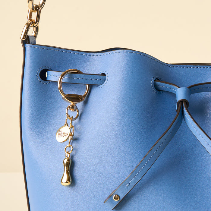 A close-up of a light blue leather bucket bag with gold hardware, a chain strap, and an I Bubble Letter Keychain Bag Charm by the-darling-effect for a unique touch.