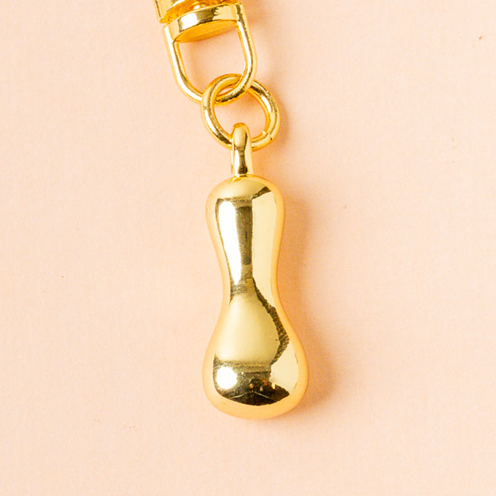 The I Bubble Letter Keychain Bag Charm by the-darling-effect features a shiny gold, bean-shaped pendant on a circular ring—an ideal personalized accessory for your bag, pictured here against a pale peach background.