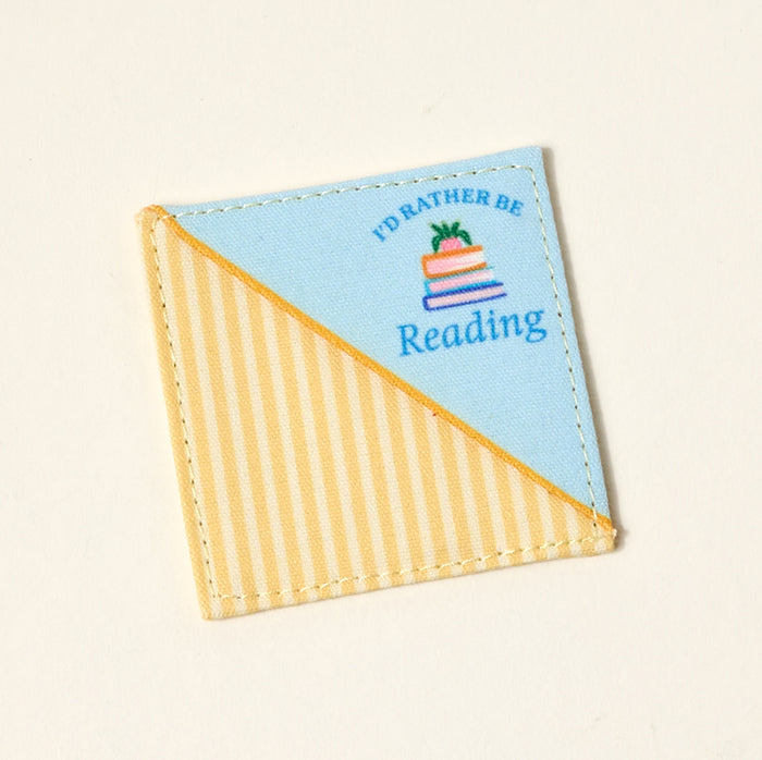 The Darling Effect’s "I'd Rather Be Reading" Corner Keeper Fabric Bookmark features diagonal yellow stripes, solid blue, a book stack, and text—perfect for book lovers.