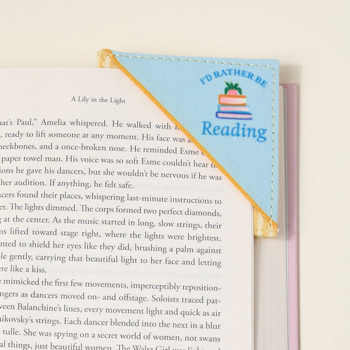 The Darling Effect’s “I’d Rather Be Reading Corner Keeper” fabric bookmark, in blue and yellow with a book stack design, clips to your book’s corner—a perfect gift for book lovers—shown resting on a white surface.