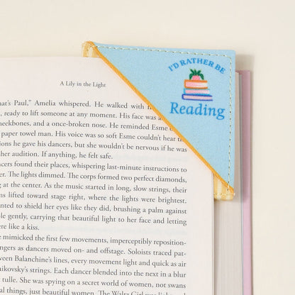 The Darling Effect’s “I’d Rather Be Reading Corner Keeper” fabric bookmark, in blue and yellow with a book stack design, clips to your book’s corner—a perfect gift for book lovers—shown resting on a white surface.