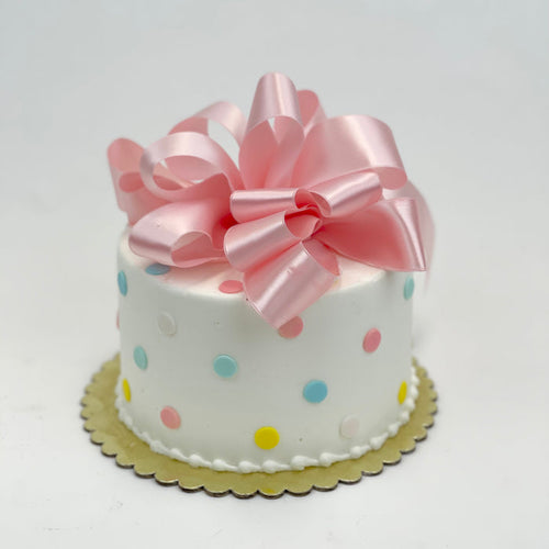 A small, single-tier cake with white fondant, pastel blue and yellow polka dots, and a pink satin bow on top.