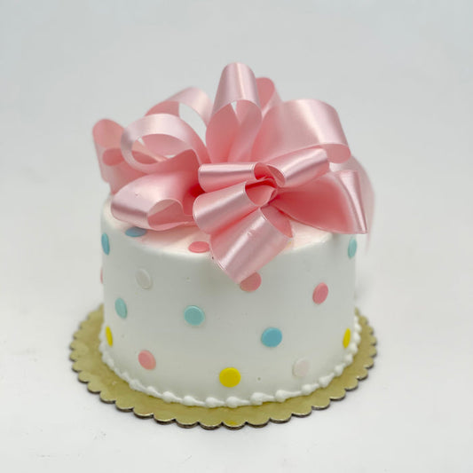 A small, single-tier cake with white fondant, pastel blue and yellow polka dots, and a pink satin bow on top.