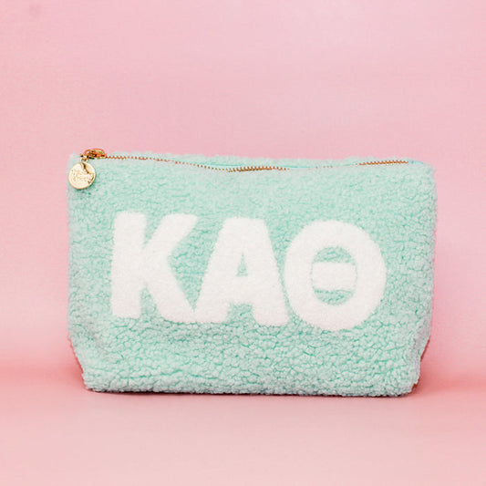 The Darling Effect’s Kappa Alpha Theta Aqua Teddy Letter Pouch features white KAΘ letters, a gold zipper, and a round charm on pastel blue textured fabric—an adorable accessory for any Theta sister.
