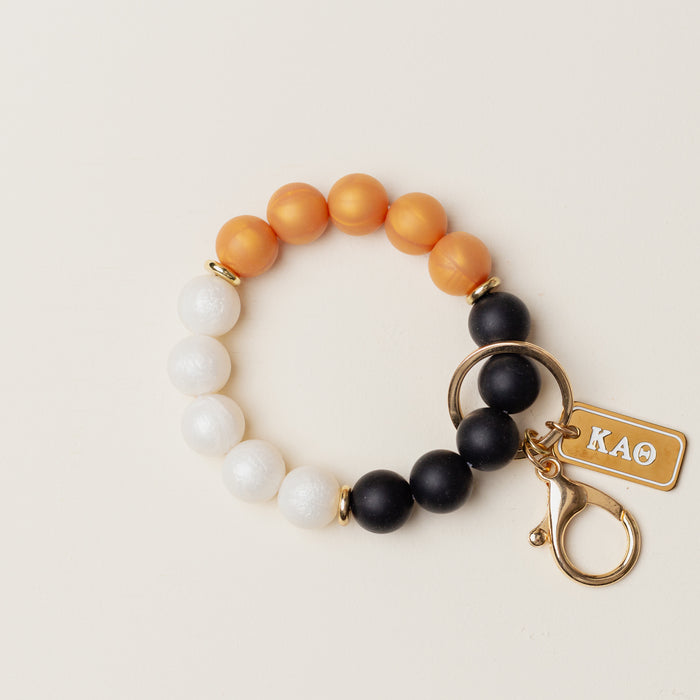 The Darling Effect’s Kappa Alpha Theta Hands-Free Keychain Wristlet features orange, white, and black silicone beads in a circle, plus a gold keyring, clasp, and engraved “KAΘ” tag.