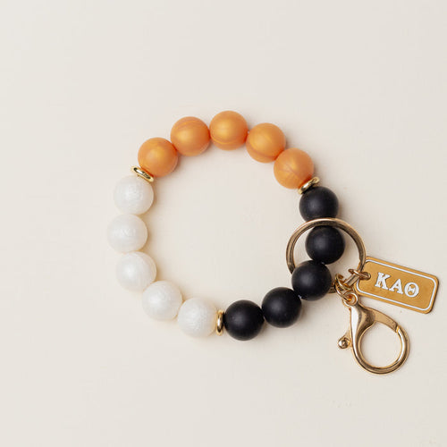 The Darling Effect’s Kappa Alpha Theta Hands-Free Keychain Wristlet features orange, white, and black silicone beads in a circle, plus a gold keyring, clasp, and engraved “KAΘ” tag.