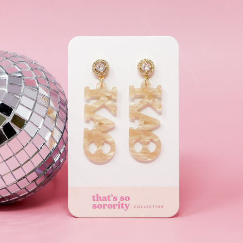 A pair of Kappa Alpha Theta Letter Gold Acrylic Earrings by The Darling Effect shown on a branded card with a silver disco ball on a pink background—perfect for any bid-day basket.