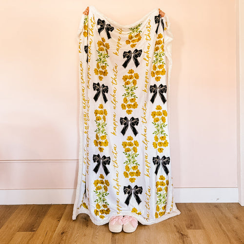 A person stands on a wooden floor, mostly hidden behind The Darling Effect's Kappa Alpha Theta Plush Sherpa Blanket featuring yellow flowers and black bows. Only their hands and pink slippers peek out from this cozy sorority blanket.