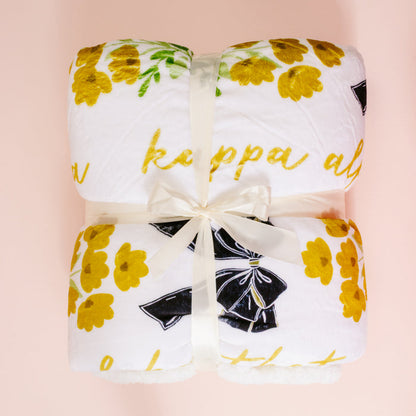 The Darling Effect's Kappa Alpha Theta Plush Sherpa Blanket—white with yellow flowers and cursive text—is neatly folded, tied with a cream ribbon, and displayed on a pale pink background, making it a cozy college dorm throw.