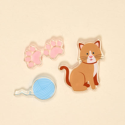 The Darling Effect's Kitty Corner Little Magnet Set features three acrylic magnets—a brown and white cat, two pink paw prints, and a blue yarn ball—perfect for adding cute kitty charm to your fridge décor.