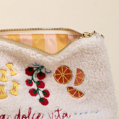 A close-up of The Darling Effect’s La Dolce Vita Teddy Pouch shows fuzzy white sherpa fabric embroidered with tomatoes, oranges, and pasta, and a partially open zipper revealing its pink and yellow striped lining.