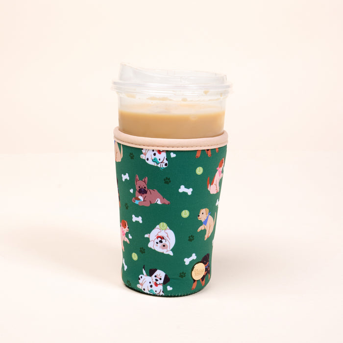 A plastic iced coffee cup with a clear lid is wrapped in The Darling Effect's Large-Dog Days Sip Savvy Drink Sleeve, featuring playful cartoon dogs, tennis balls, and bones on a plain, light background.