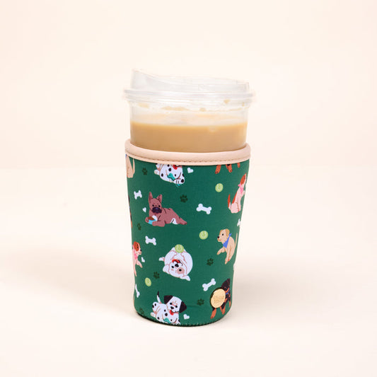 A plastic iced coffee cup with a clear lid is wrapped in The Darling Effect's Large-Dog Days Sip Savvy Drink Sleeve, featuring playful cartoon dogs, tennis balls, and bones on a plain, light background.