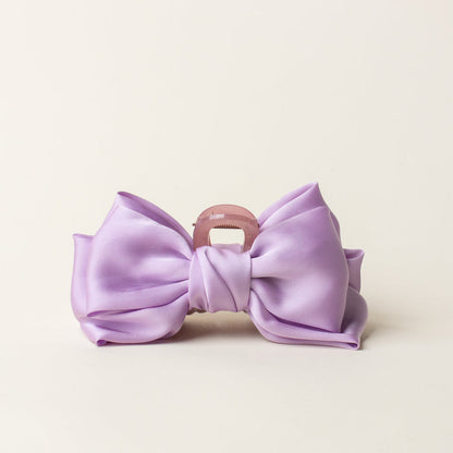 The Lilac Satin Bow Claw Clip by the-darling-effect features a large lavender satin bow on a light pink clip, adding charm to your updos. Shown against a simple off-white background.