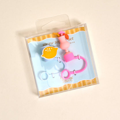 The Darling Effect’s Limoncello Love Straw Topper Set includes leak-resistant pink and yellow lemon-shaped and pink ring silicone toppers, adding fun to your drinkware accessories. Displayed on a light blue and yellow striped background.