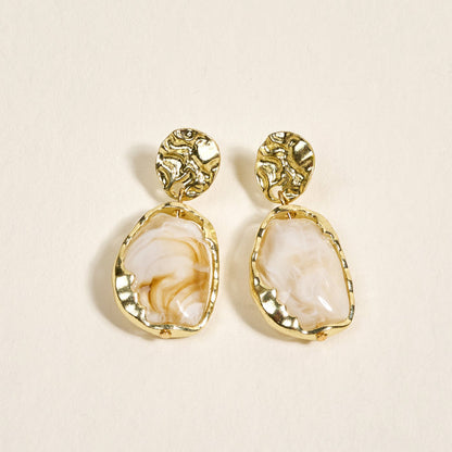 The Darling Effect's Liquid Gold Standout Earrings feature round textured gold studs and irregular white marbled stones with a high-shine finish, displayed on a light cream background.