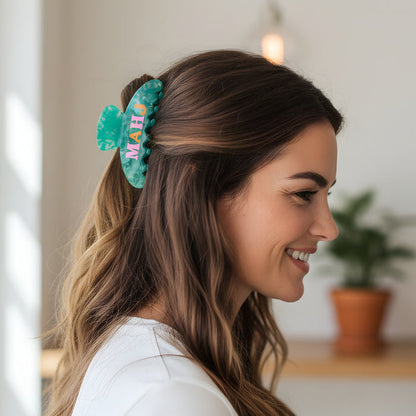 A woman with long, wavy brown hair smiles in profile, wearing The Darling Effect's Mahj Word Up Claw Clip in green. A potted plant and a hanging light are softly blurred in the background.