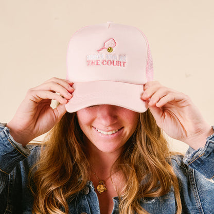 A person smiles, holding the brim of the-darling-effect’s Meet Me at the Court Trucker Hat—pink with "meet me at THE COURT," a mini tennis racket and ball—stylishly paired with a denim jacket.