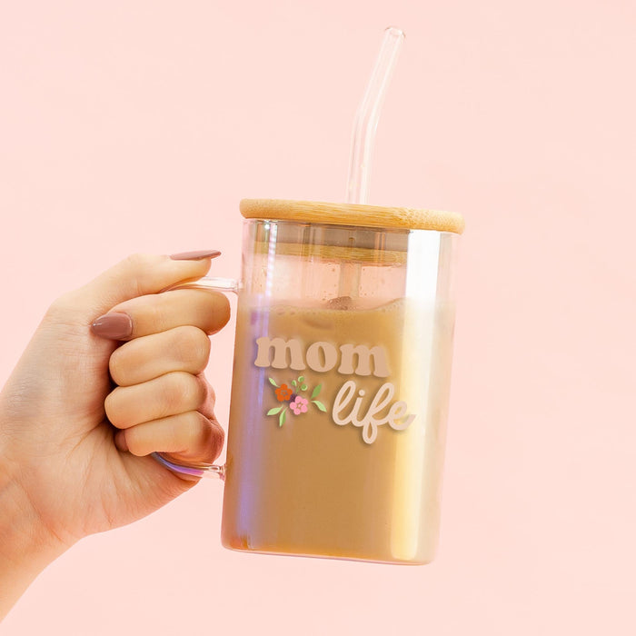 A manicured hand holds the-darling-effect's Mom Life Square Glass Cup, featuring a floral design, bamboo lid, and clear straw, set against a light pink background.