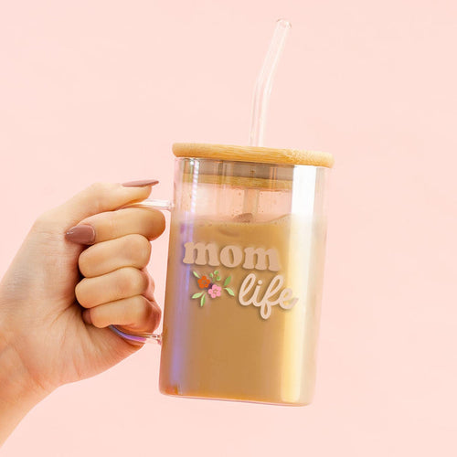 A manicured hand holds the-darling-effect's Mom Life Square Glass Cup, featuring a floral design, bamboo lid, and clear straw, set against a light pink background.