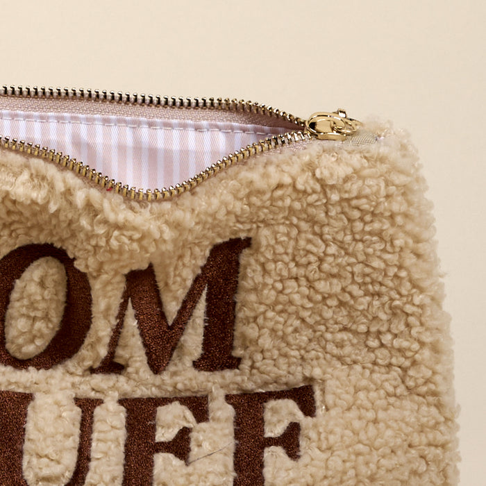 A close-up of The Darling Effect's Mom Stuff Teddy Pouch shows its beige sherpa fabric with brown "Mom Stuff" embroidery and an open zipper revealing a white and light pink striped lining inside.