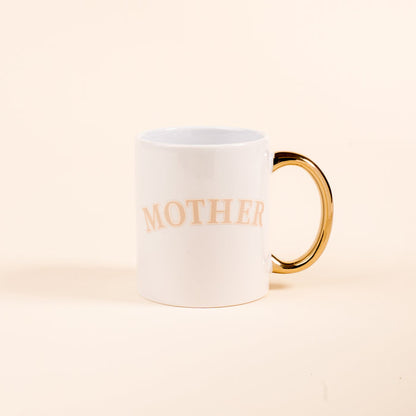 The Darling Effect’s Mother 11 oz Cozy Cute Ceramic Mug features a white design with a golden handle and “MOTHER” in light beige letters—perfect for coffee or tea. Photographed on a plain light beige background.