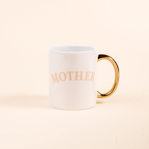 The Darling Effect’s Mother 11 oz Cozy Cute Ceramic Mug features a white design with a golden handle and “MOTHER” in light beige letters—perfect for coffee or tea. Photographed on a plain light beige background.