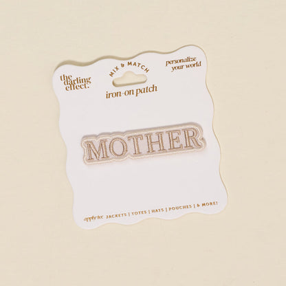 The Darling Effect’s Mother Mix & Match Iron-On Patch features "MOTHER" in gold on white, attached to a wavy-edged gold-accented backing—ideal for customizing jackets, totes, hats, and more. Discover endless style with our iron-on patches!.