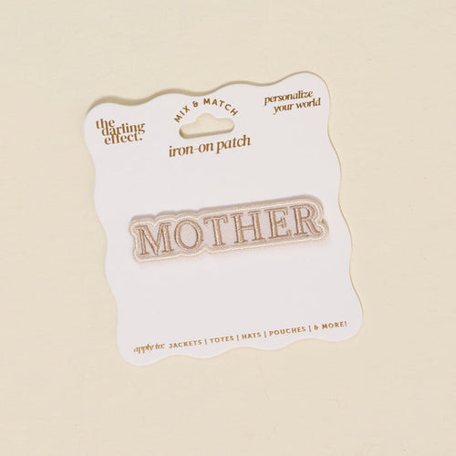 The Darling Effect’s Mother Mix & Match Iron-On Patch features "MOTHER" in gold on white, attached to a wavy-edged gold-accented backing—ideal for customizing jackets, totes, hats, and more. Discover endless style with our iron-on patches!.