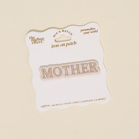 The Darling Effect’s Mother Mix & Match Iron-On Patch features "MOTHER" in gold on white, attached to a wavy-edged gold-accented backing—ideal for customizing jackets, totes, hats, and more. Discover endless style with our iron-on patches!.